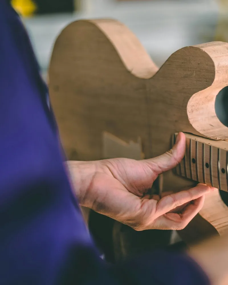 Manos sosteniendo un cuerpo de guitarra en madera desnuda junto al mástil con el diapasón trasteado, en un banco de trabajo